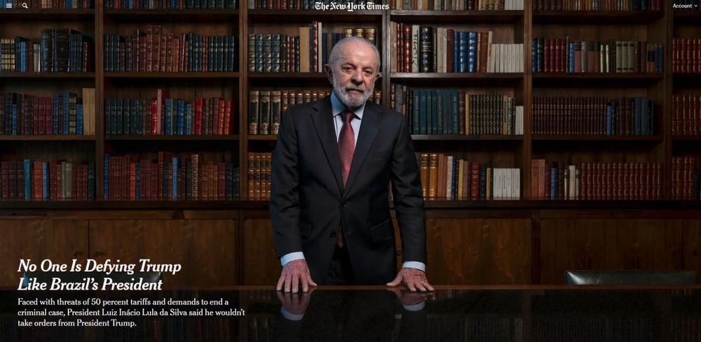 Foto publicada pelo The New York Times mostra Lula em pé atrás de uma mesa escura, com as duas mãos apoiadas no tampo brilhante, em pose firme e imponente. Ele veste um terno escuro elegante, camisa azul clara e gravata vermelha com estampa discreta. Ao fundo, há uma estante de madeira preenchida por livros de lombadas coloridas, em tons de vermelho, azul, verde e dourado, compondo um cenário que lembra biblioteca de filme sobre espiões ou julgamento internacional.

O clima da foto é todo montado pra passar autoridade, experiência e aquele ar de “líder que não baixa a cabeça”, bem condizente com o título que aparece no canto inferior esquerdo: “No one is defying Trump like Brazil’s president” (Ninguém está enfrentando Trump como o presidente do Brasil). Abaixo, o subtítulo reforça a ideia de confronto direto, dizendo que mesmo com ameaças de tarifas de 50% e pressões diplomáticas, Lula se recusou a obedecer ordens de Trump.
