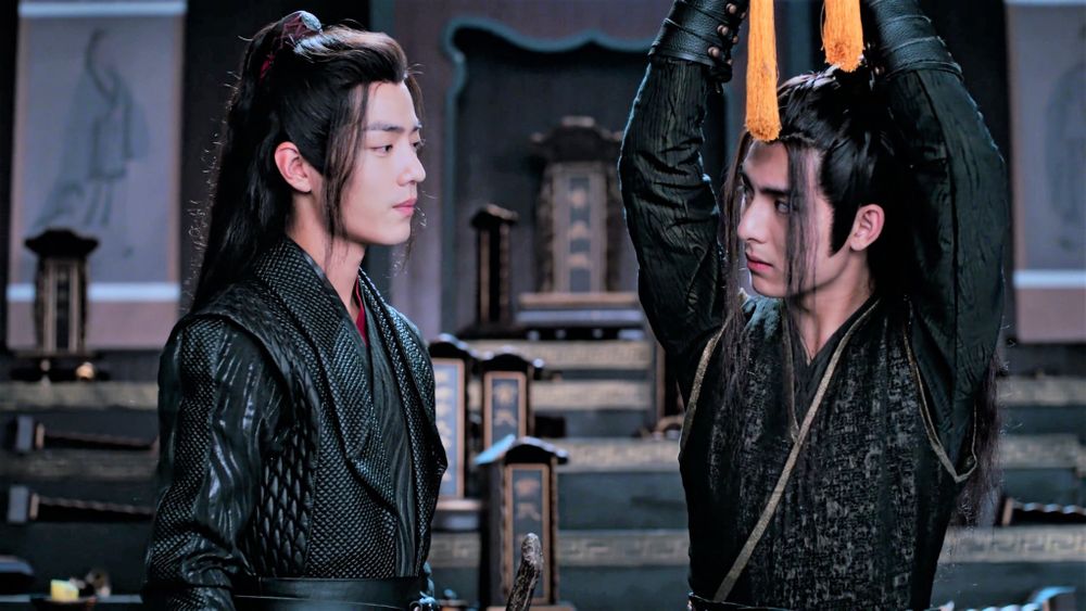Wei Wuxian cautiously regards Xue Yang while Xue Yang looks back at Wuxian with this high slightly tilted to the side, waiting for him to proceed. Xue Yang's hands are bound above his head, just out of frame, though the tassels at the end of the rope used dangle back into frame over his head.
