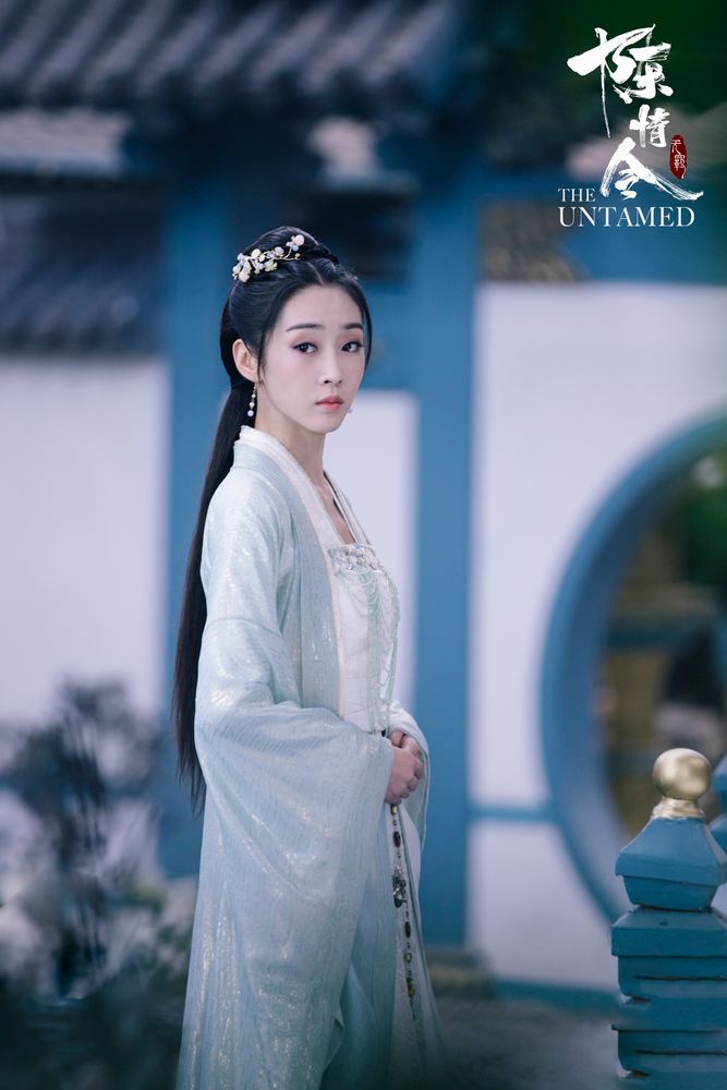 Jiang Yanli stands in profile, head turned toward the viewer, directing a solemn expression at an offscreen target. Her robes are pale, something ice blue over white, and a floral hairpiece encircles halfway around the bun holding some of her hair. Her surroundings appear to be a courtyard, a blue rail post topped with gold in front of her, some indistinct plant life behind her, and in the furthest background is a sloped tile roof surmounting a white wall through which there is a blue trimmed circular archway.