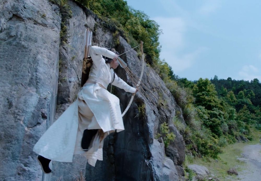 Jin Ling leaps through the air alongside a cliff with an arrow nocked to his bow and the bow drawn, aiming at an out of frame target.