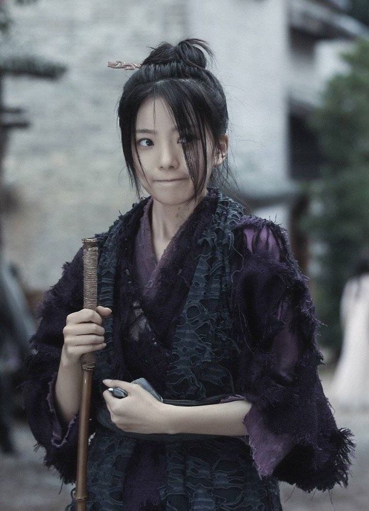 Image features A-Qing, shown from hips up. She holds a long walking stick in her right hand, the top of which is level with her shoulder, and a money pouch is clenched in her left hand against her waist. Her lips are pressed together such that her mouth forms a flat line but still hints at a smile as her eyes are looking sideways beyond the right side of the image, giving her an amused but considering air.