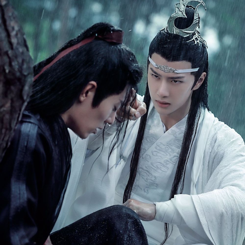 Wei Wuxian and Lan Wangji are outside in the rain. Wei Wuxian is seated against a tree, slightly hunched over, with his eyes closed. Lan Wangji kneels facing him, right hand clenched level with his lower chest and right hand with only his pointer and middle fingers extended and touching Wuxian's forehead, transferring spiritual energy, face concerned but determined.