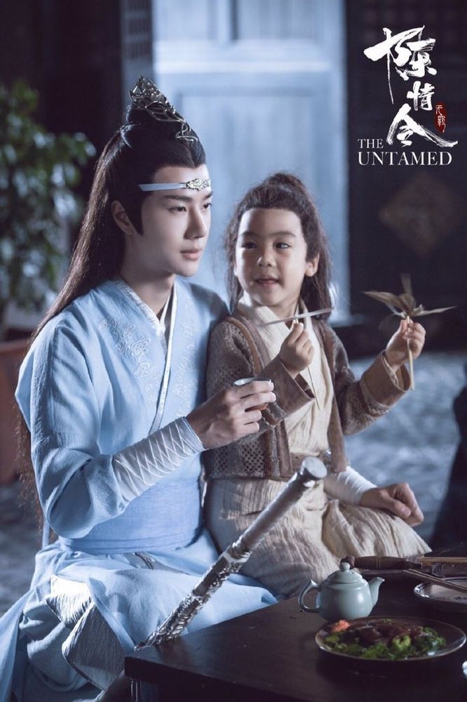 Lan Wangji contentedly sits at a table set with dishes and a teapot in a restaurant in Yiling with A-Yuan sitting on his left leg. A cup of tea is held in his left hand suspended mid-way between the table and taking a drink while his left hand rests on A-Yuan's knee. A-Yuan has turned his head to look at Wangji, each of his hands holding one of a pair of woven grass toys attached to sticks. Bichen has been set down to Wangji's right, leaning against the table so that the hilt plus an equal amount of sheath are visible above the tabletop.