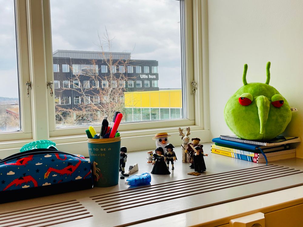 A deep window shelf with an eclectic display of figurines, a number of which are Playmobil: a hockey player, two 17th century figures, a monk, Martin Luther, Olaf and his destiny friend from the movie Frozen and a moose. There is also a green plush invasive bug with red eyes, a pencil case covered with fire-breathing dragons, and a cup that says “time to be a unicorn” with a unicorn riding a bike serving as a pencil holder. 