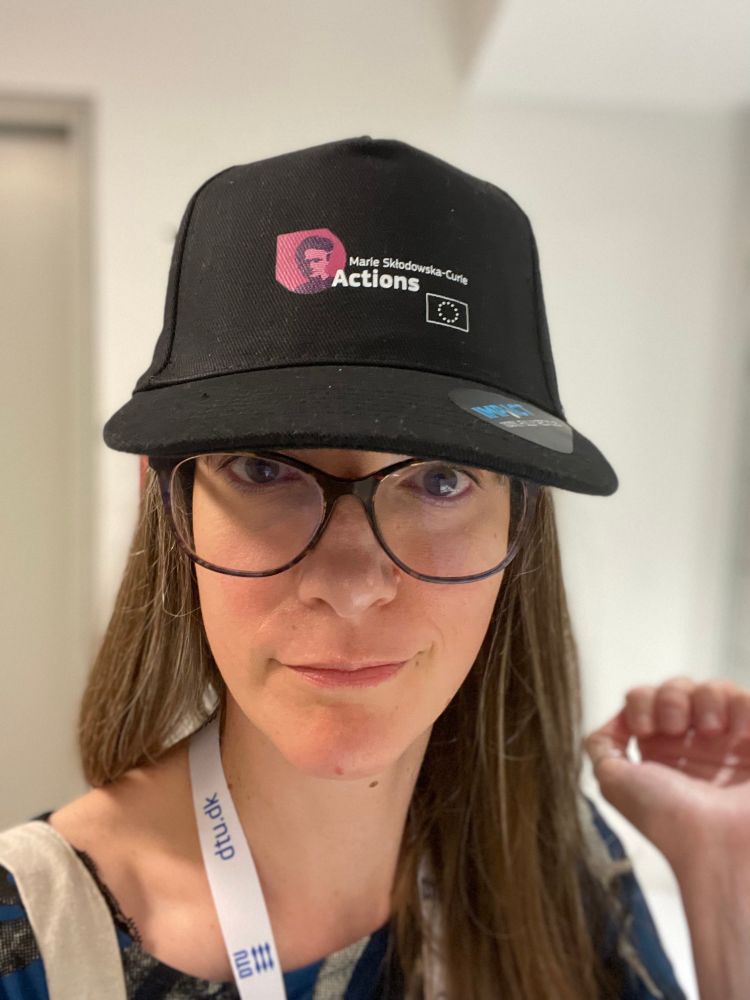 A white woman with long brown hair and big tortoiseshell glasses models a black baseball cap with the Marie Skłodowska-Curie Actions and EU logos. 