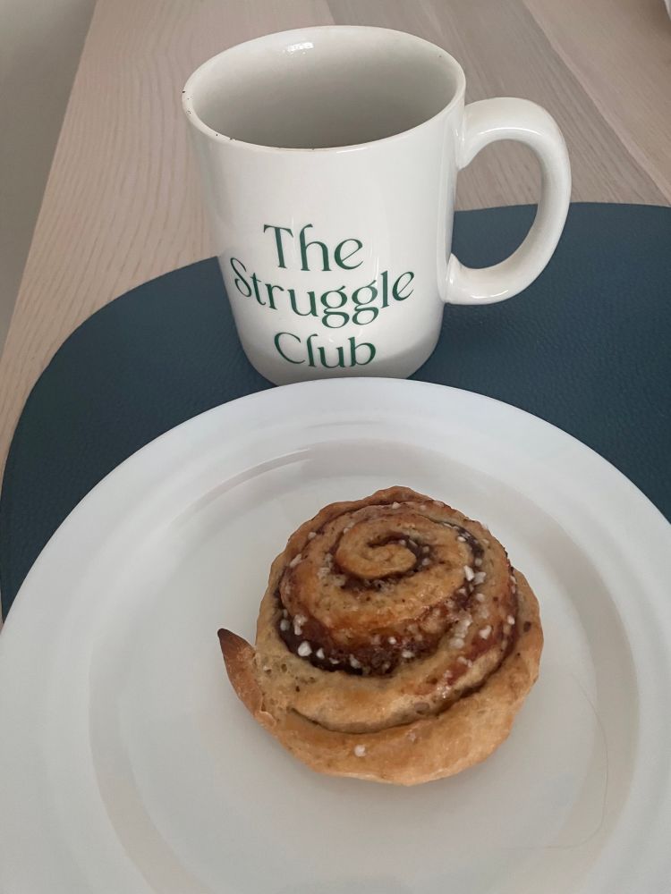A small gluten- and lactose-free cinnamon bun on white plate. Behind the plate is a white mug that says “The Struggle Club” in green serif lettering. The mug contains very strong coffee. Both the mug and the plate are on a dusky blue placemat on a wooden dining table. This was for the picture. I ate the pastry while sitting on the sofa. 