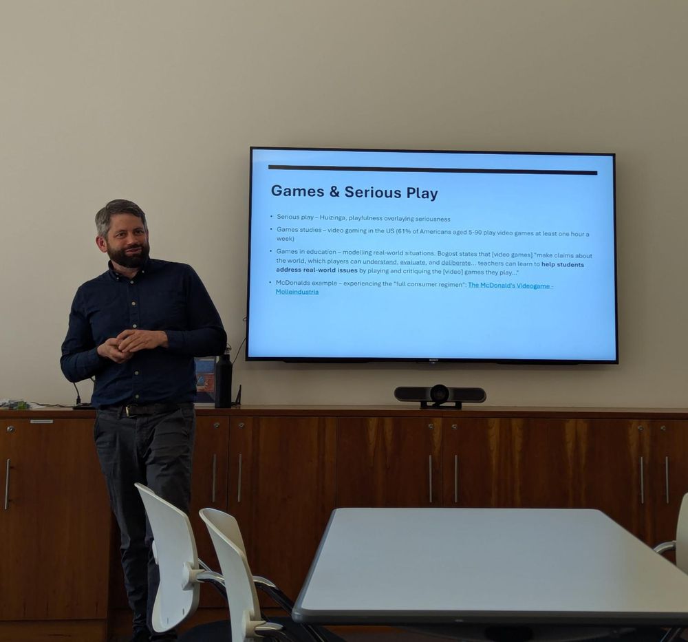 A presenter stands at the front of the room. Text on the presentation slide discusses games and serious play. 