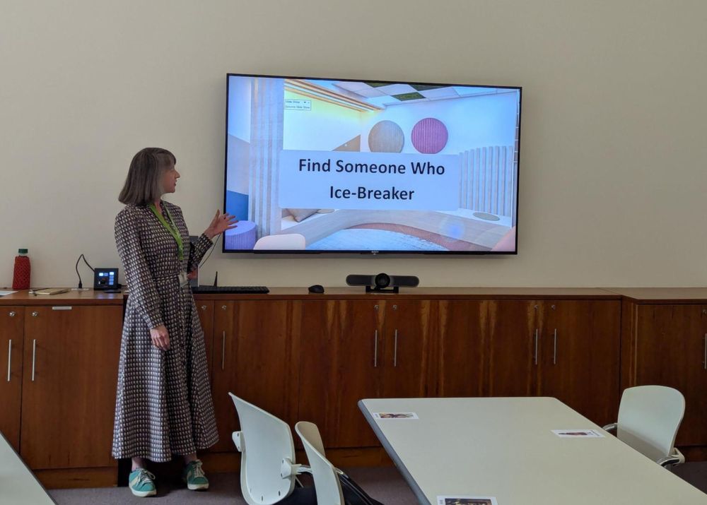 A presenter at the front of a room. The title of the presentation slide on the television screen reads Find Someone Who ice-breaker. 