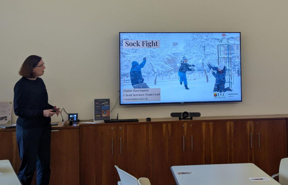 A presenter at the front of a room. The title of the presentation slide on the television screen reads Sock Fight.