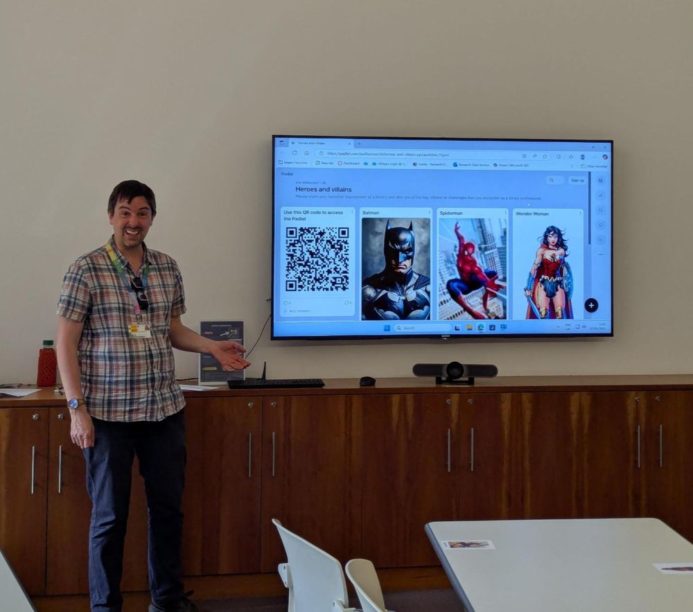 A presenter at the front of a room. The television screen shows a padlet with various superheroes. 