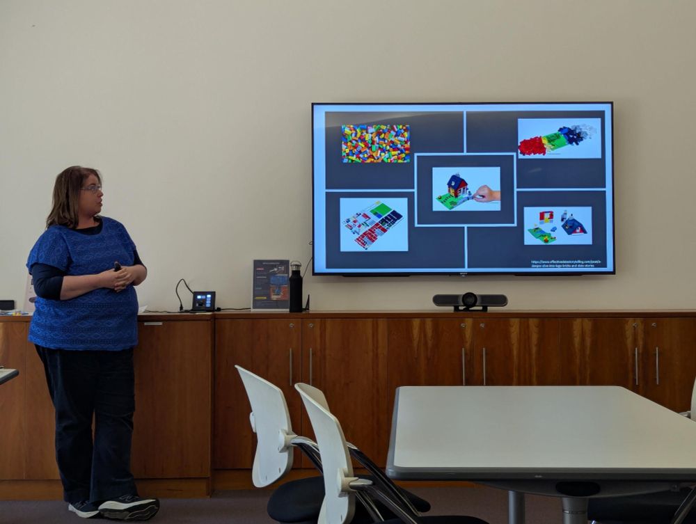 A presenter stands at the front of a room. Images on the television screen show Lego and the process of using it to build a house. 