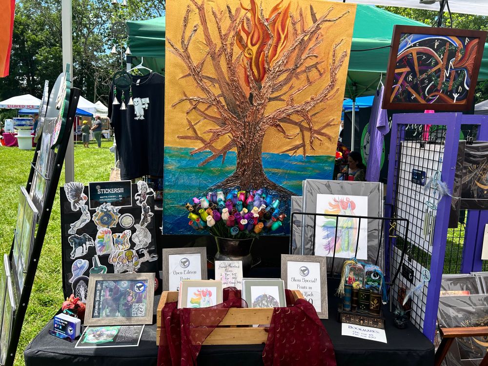 A heavily textured painting of a tree with a gold background hangs over a table with a black tablecloth. On the table, from left to right: a black display with fantasy creature stickers, a stack of business cards, a wooden crate full of small art prints, a silver vase full of multicolored roses, a metal rack with larger art prints, and a small book-shaped wooden display full of tasseled bookmarks. 