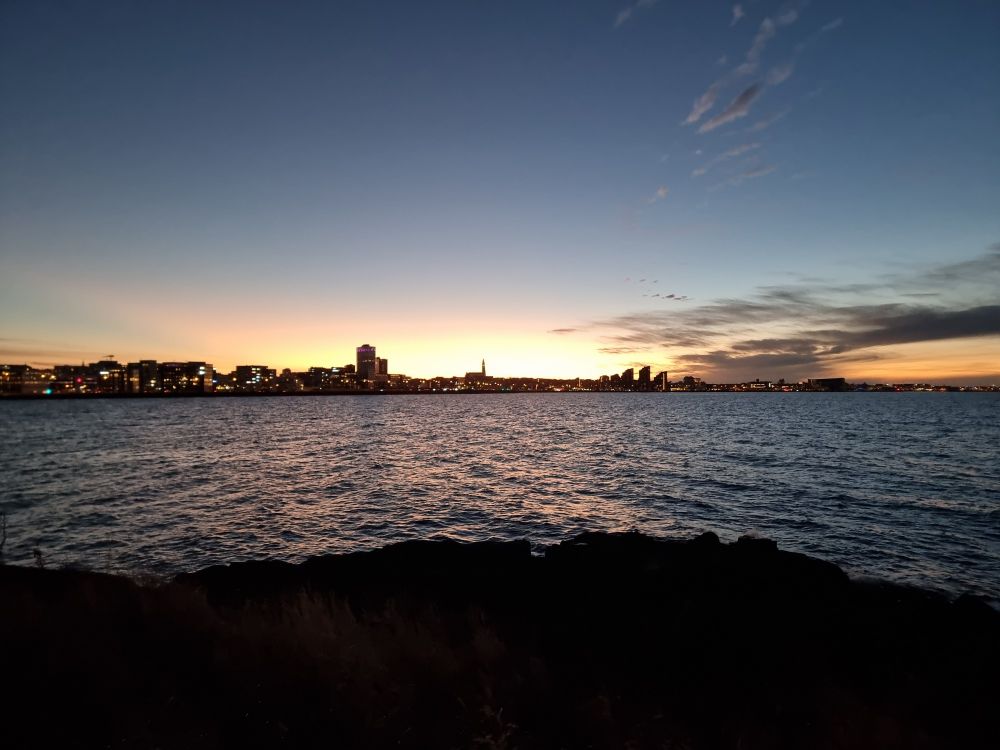 Skyline of Reykjavík, Iceland that was shot a few days ago. In the front is the shore, overlooking the sea towards a city in the distance. Behind the sun is going down  casting yellow and orange across the sky.