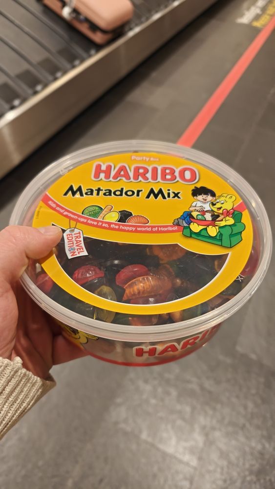 A 1kg tub of Haribo Matador Mix containing a mix of gummi sweets and licorice, in front of an airport luggage conveyor belt