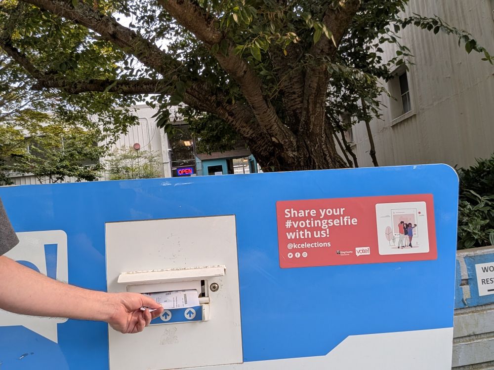 A ballot being dropped into a blue and white outdoor ballot box. In the background, trees are in full summer leaf.