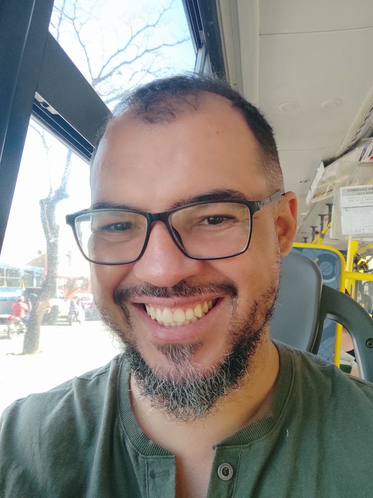 Homem de cabelo preto e barba preta com fios brancos. Usa óculos de armação preta. Ele está sorrindo. Ao fundo, aparece o interior de um ônibus urbano. 