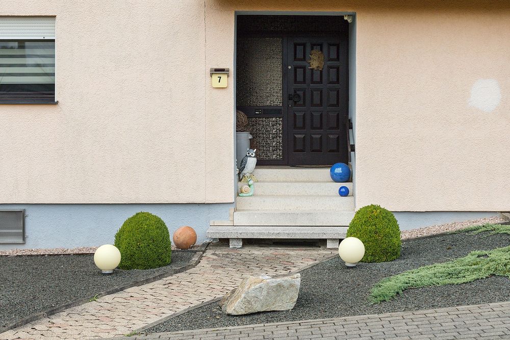 Een voordeur, met een beige huis. Nummer 7. Een beeld van een uil en een slak op het trapte naar de deur, verder een pad tussen de donkergrijze steentjes. een grote steen, 2 bosjes, rond geknipt en een assorti aan stenen bollen, in de kleur geel, blauw en roodachtig.