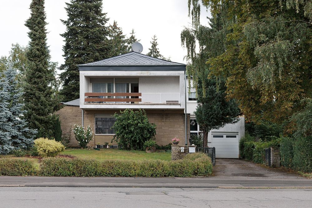 Een vrijstaand huis tussen de bomen, met wat elementen die ik thuis minder zie, naaldbomen, leistenen dak. Er is een balkon en de deur is open.