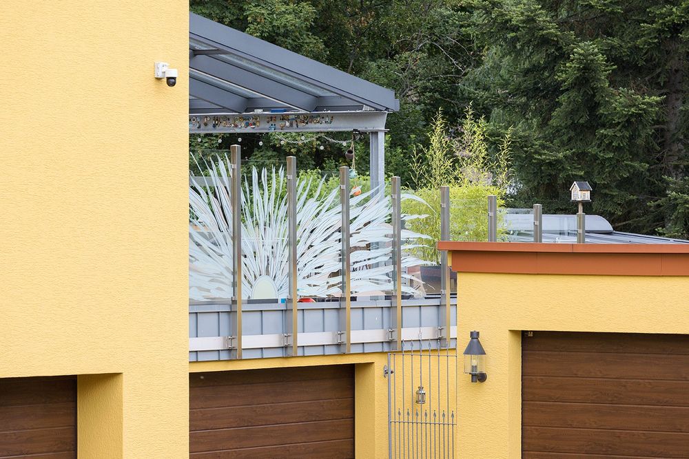 Een geel huis, maar het middelpunt is een metaal en glas 'schutting'met een hysterische zon. Je ziet ook een camera, een vogelhuisje in de vorm van een huis en op de achtergrond bos.