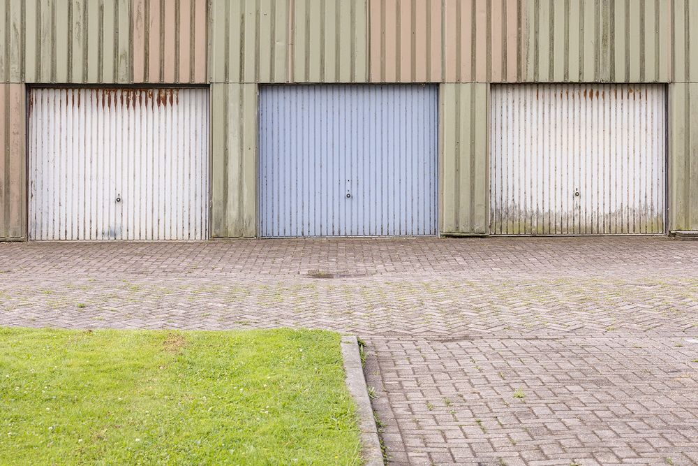 3 metalen garages, met deuren in beige en lichtblauw, de omlijsting is groen metaal, ervoor straat en een hoek met een grasveld.