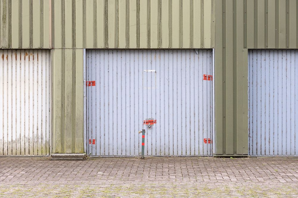 Lichtblauwe garage, lichtblauw, de omlijsting is groen metaal, ervoor straat. Voor de deur een paaltje en de rode stickers, waar je het woord 'verzegeld' nog op ziet staan.