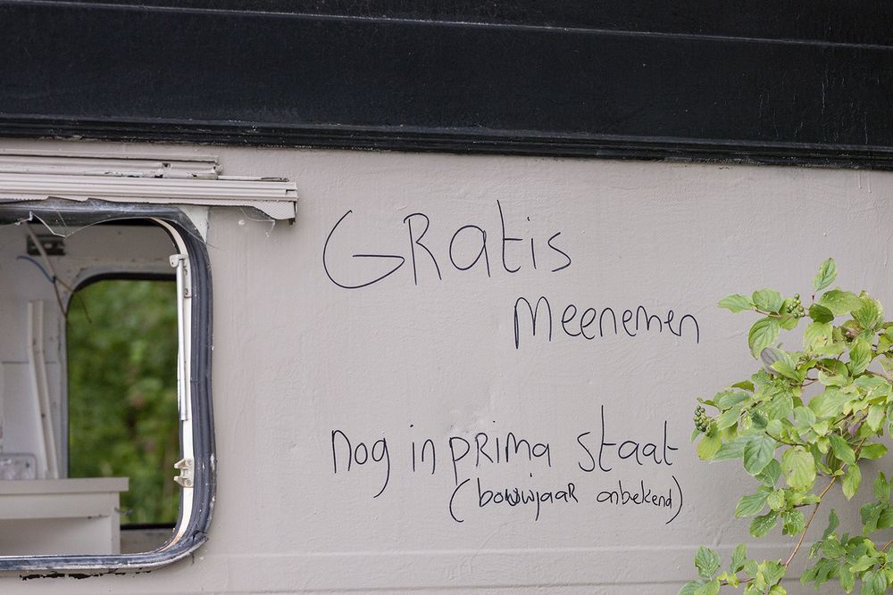 Een kapotte grijszwarte caravan, met de tekst: Gratis meenemen, nog in prima staat (bouwjaar onbekend).