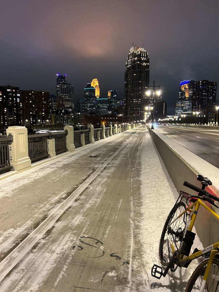 Illuminated Minneapolis skyline at night from the Central Avenue bridge. A yellow and white bike is propped against the concrete barrier dividing the car lanes from the bike lane/sidewalk. The bridge has a dusting of snow on it. It’s overcast, so the light from the buildings can be seen in the sky.