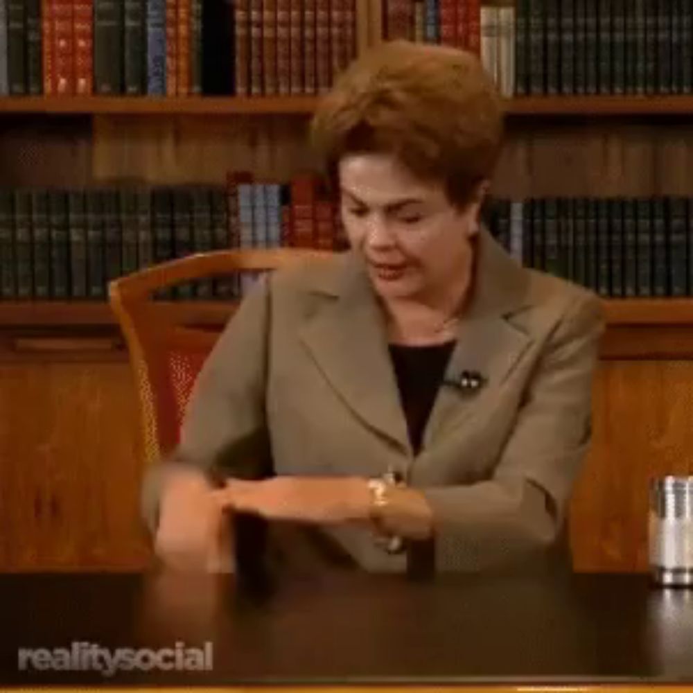 a woman in a suit is sitting at a desk in front of a bookshelf with realitysocial written on it