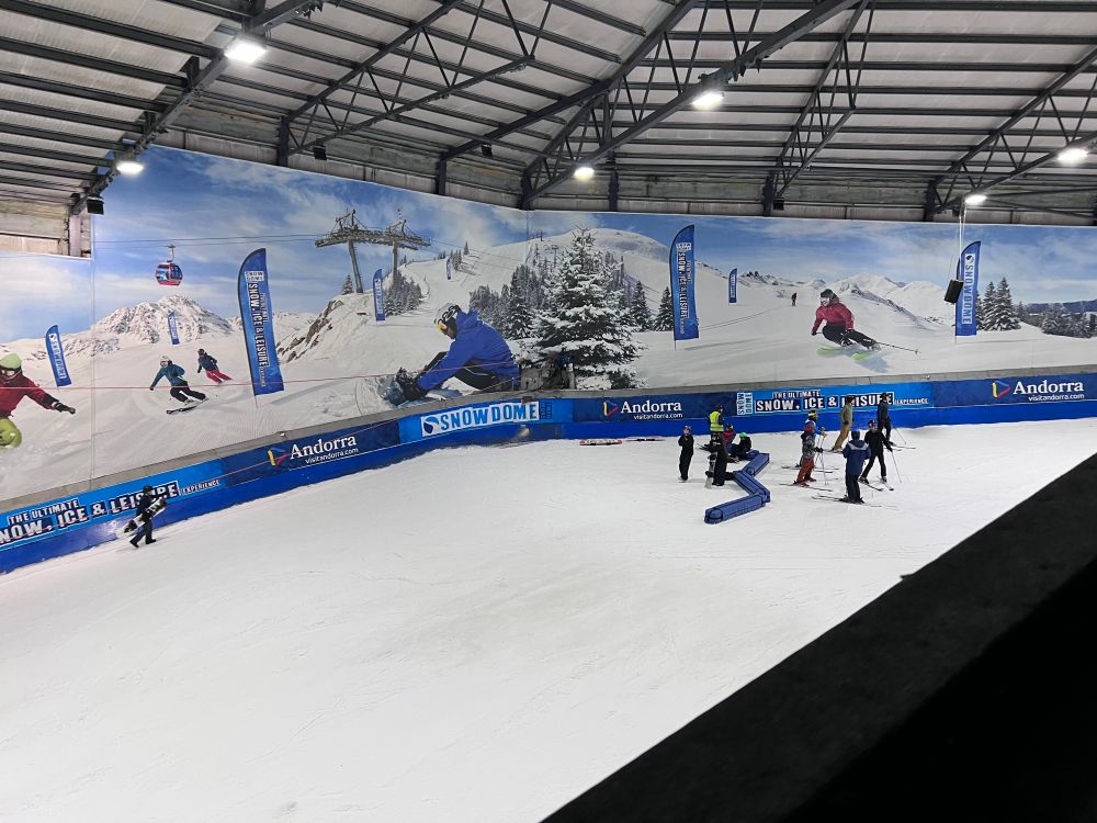 The bend on an indoor ski slope, with people standing behind blue barriers on the apex of the corner. There’s a long poster on the wall that shows people skiing and snowboarding down a mountain, with advertisements for the ‘Snowdome’, and Andorra.