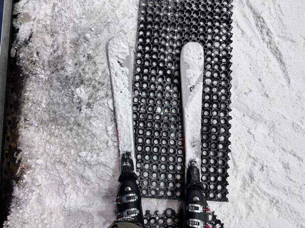 A pair of skis standing on a rubber, latticed matting. Snow covers the wording. There are a pair of booted feet attached.
