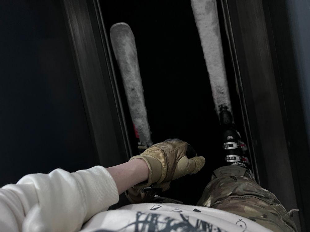 Looking down at snow-covered skis, from about chest-level. The skis are attached to boots, which are attached to a pair of legs wearing camo trousers. The photographer’s wearing a white Sleep Token hoodie, though the graphic is barely visible.