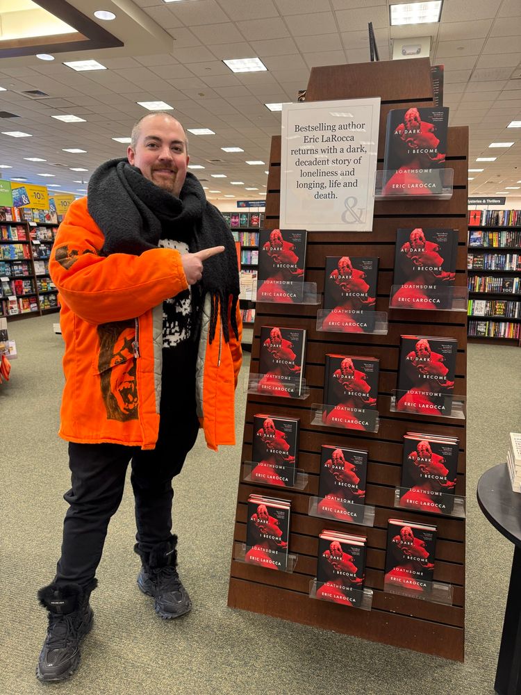 Eric in an orange jacket and standing beside an end cap filled to the brim with hardcover copies of AT DARK, I BECOME LOATHSOME