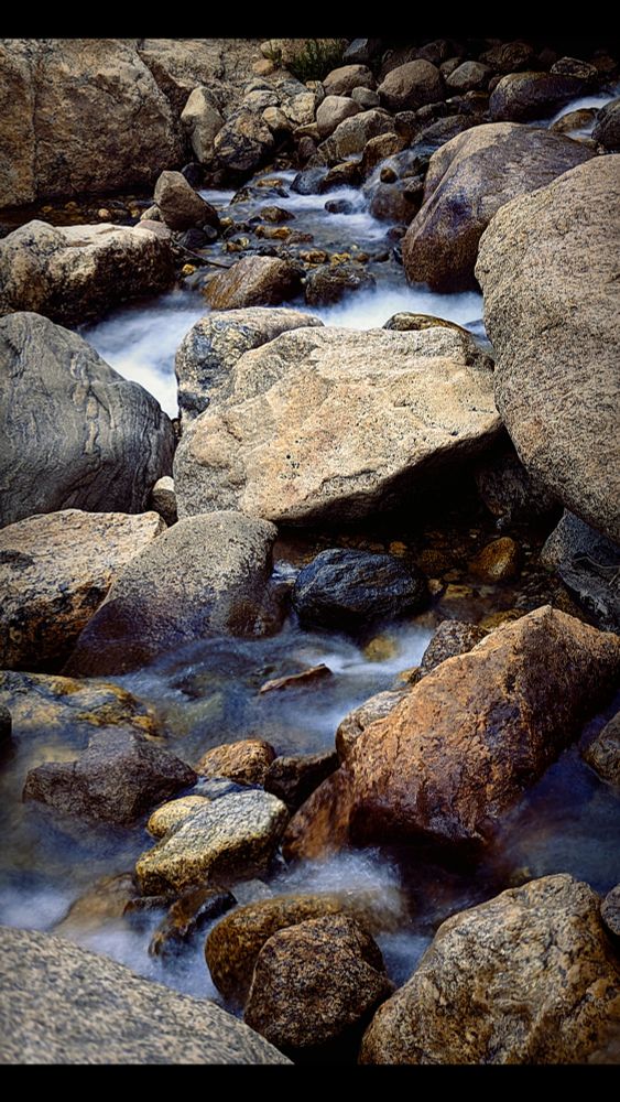 Long exposure shot of rushing water flowing right to left along rocks. The rocks are mostly underwater, with only a bit of them poking out of the current. Colors are warm on the rocks, but deceptively cool as you get closer to the water.