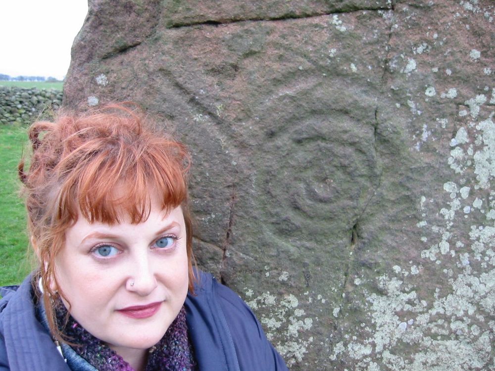 A photo of a young white woman with red hair in a blue anorak posing in front of a lichen covered standing stone etched with a spiral. Her expression is thoughtful and and maybe a bit surprised. 