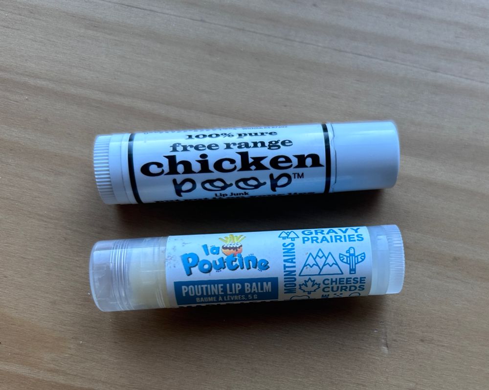 Two tubes of lip balm lying horizontally on a wooden surface. The upper tube is white with black writing and reads: 
100% Pure
free range
Chicken 
Poop

The lower tube is transparent with the yellowish balm inside showing through at one end, and a white label with blue writing:
La Poutine
Poutine Lip Balm
Baume à lèvres, 5g

And on the right side of the label, some stylized drawings of mountains, a totem pole, a maple leaf, and the words:
GRAVY
PRAIRIES
MOUNTAINS
CHEESE CURDS
