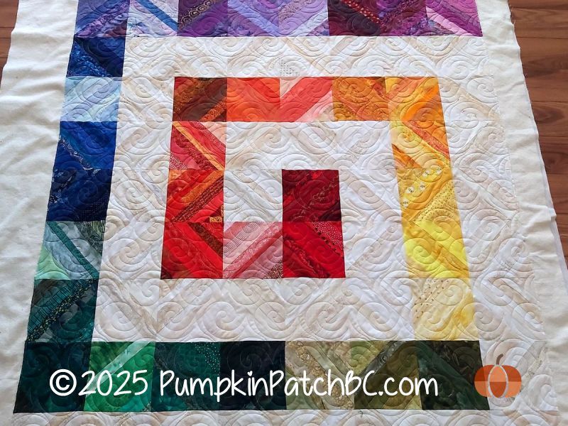 A patchwork quilt with blocks pieced from leftover strips, arranged in a colorful spiral that resembles a rainbow, from red to yellow to green to blue to purple, with white and cream squares in between