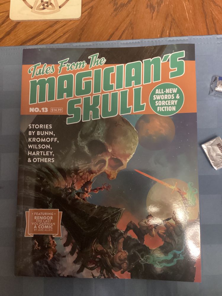 The cover of Tales From the Magician’s Skull #13. In the background looms a massive skeleton. A giant sword pierces its chest. A horse of strange creatures with spikes and glowing eyes are crawling down the sword towards the party of magical adventurers in the foreground, who have drawn swords and created spells to defend themselves.
