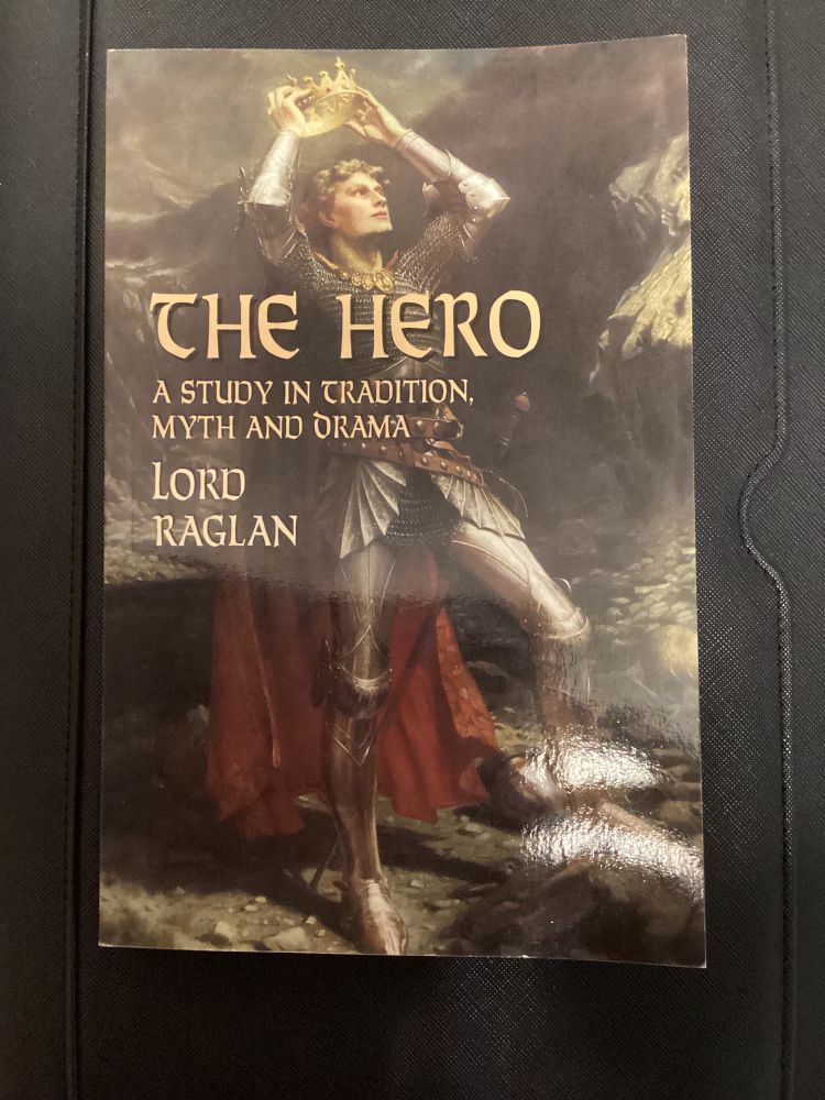 The cover of The Hero by Lord Raglan. A knight in full armor and a red cloak stands with one foot braced on a stone. He is holding a crown over his own head.