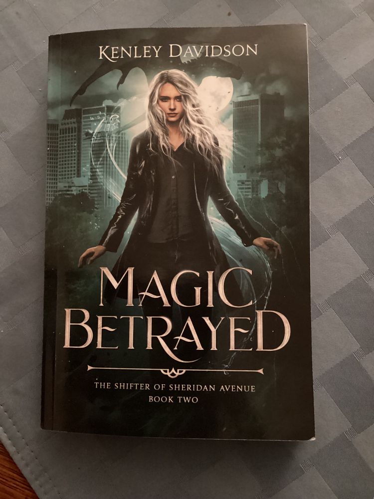 The cover of Magic Betrayed by Kenley Davidson. A woman with long, white-blonde hair stands in the foreground. She is dressed in black leather and cotton. Behind her stand tall skyscrapers. A large black dragon is flying through the night sky, framed by a full moon.
