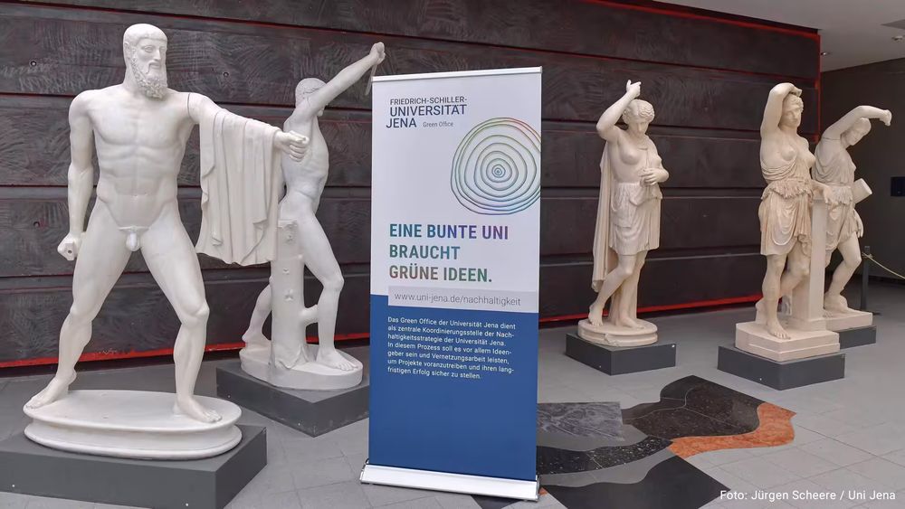 Im Foyer im Campusgebäude der Uni Jena stehen weiße Skulpuren und ein Aufsteller mit dem Text "Eine bunte Uni braucht grüne Ideen". Foto: Jürgen Scheere / Uni Jena