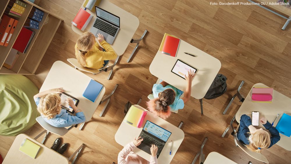 Several children are sitting at individual desks in a classroom, working on laptops, tablets or smartphones.