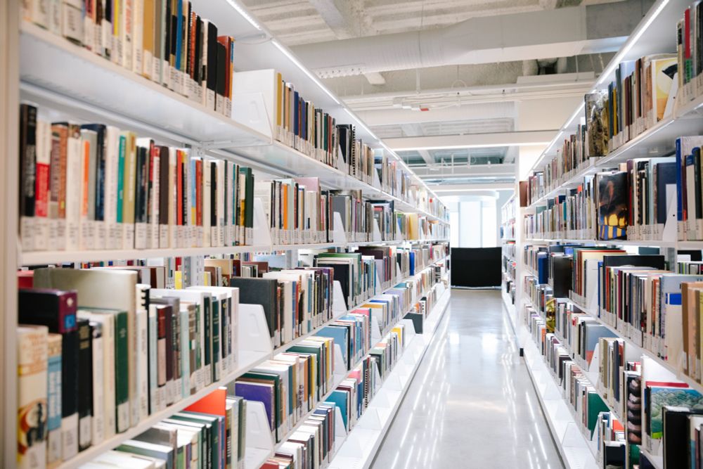 A photo gazing down the walkway between two of the library's bookshelves towards an open, bright window.