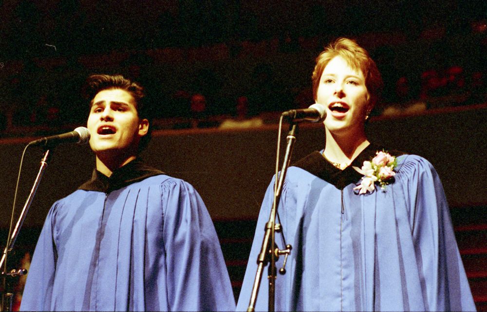 Two individuals in blue graduation gowns and black stoles, singing at a microphone during a ceremony.