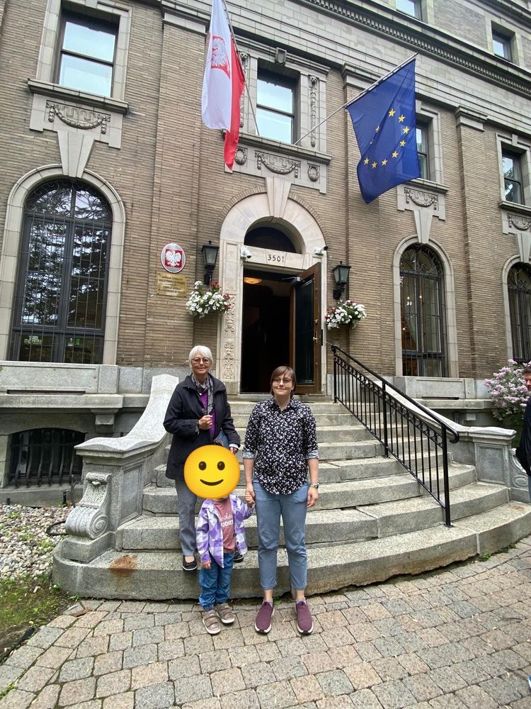 Two women and a toddler on the steps standing in front of a building with two flags (one Polish and one European Union) 