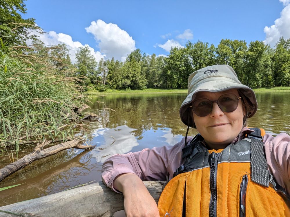A happy woman, wearing an orange life vest and bucket hat, leaning on a log on a river with trees in the background.