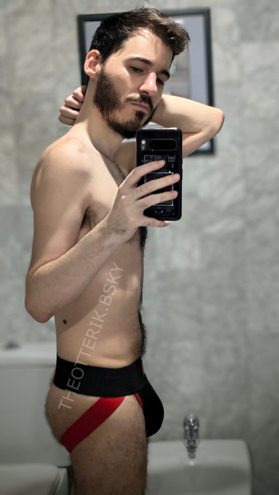 Bathroom mirror selfie of a white hairy man just wearing a black and red jockstrap. The picture is from the side, revealing ass side and bulge.