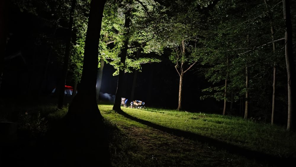 Photograph of a campsite at night. There is a super bright floodlight being blocked by a tree in the foreground, the light makes the ground look like daytime