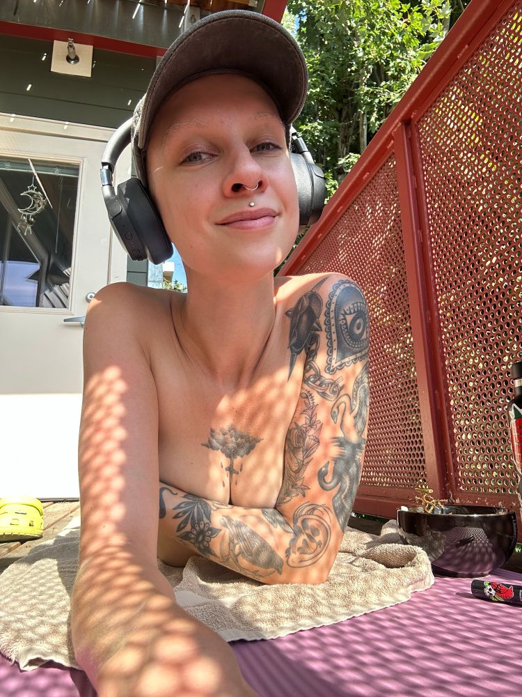 Pepper is sunbathing on their stomach, their tattooed arm concealing their chest and a sly smile on their beautiful face