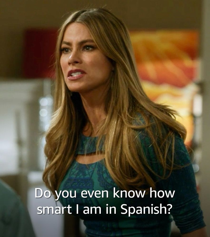 The image shows Gloria, a character from Modern family, with long brown hair and a serious facial expression. She is wearing a patterned blue-green top and appears to be in a conversation or argument. The text overlaid on the image says, “Do you even know how smart I am in Spanish?”