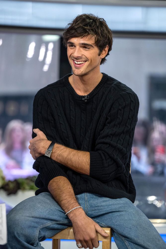 Jacob Elordi is wearing a black sweater, jeans, and some accessories like a watch, bracelet, and necklace.